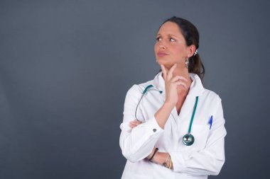 Gri arka planda, elleri çenesinin altında izole edilmiş, şüpheli ve şüpheci bir ifadeyle yana bakan zarif Avrupalı genç bir doktor portresi. Gri arka planda kapalı duruyor..