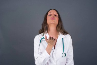 Sevimli çekici doktor kadın kameraya öpücük yolluyor, sevgilisine aşkını gösteriyor ya da uzaktan veda ediyor, gri stüdyo arka planında izole bir şekilde..