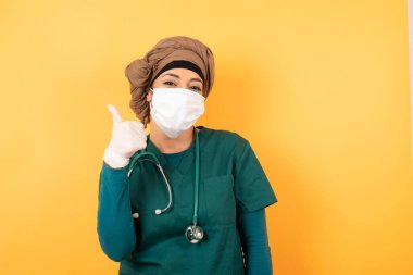İyi iş! Yeşil tıp üniformalı güzel Müslüman kadın portresi dışarıda dikilirken başparmak hareketi yapıyor. Olumlu insani duygu yüz ifadesi vücut dili. Komik kız.