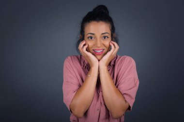 Hayalperest genç kadın ellerini çenesinin altına bastırıyor, mutlu bir ifadeyle bakıyor, dişlek gülüşü var, gri arka planda soyutlanmış.