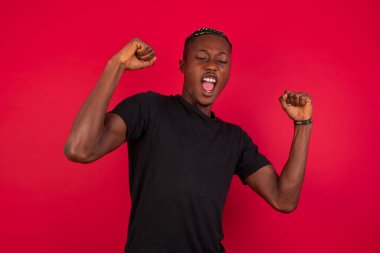 Evet, ben kazandım. Kırmızı arka plan öğrencisinin üzerine siyah tişört giyen Afro-Amerikan bir erkeğin portresi zafer ve zaferle yumruğunu kaldırıyor. Mutluluktan sırıtarak başarı elde ediyor..