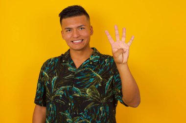 Genç İspanyol adam gri duvarın önünde duruyor. Dört numaralı parmaklarıyla işaret ediyor ve gülümsüyor. Kendinden emin ve mutlu..