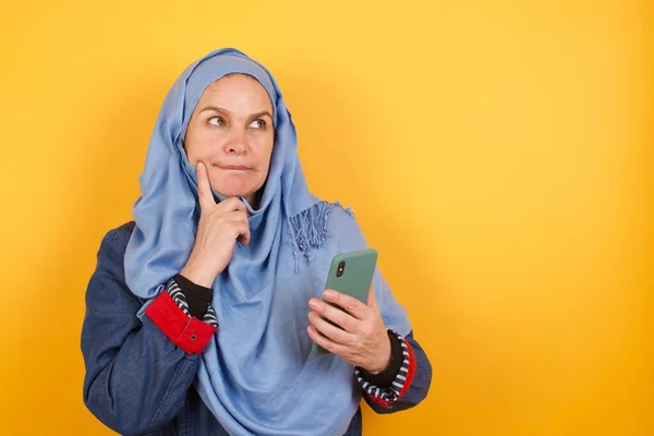携帯電話を使用して壁の背景に隔離されたヒジャブの中年のイスラム教徒の女性を夢見る考えのイメージ — ストック写真