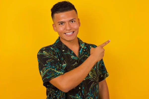 Handsome man pointing away and smiling to you while standing against yellow background.