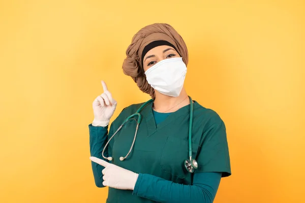 ポジティブな表情を持つ緑色の医学服の若い美しいイスラム教徒の医師の女性は あなたのプロモーションテキストや広告のための空白のコピースペースで指で示しています — ストック写真