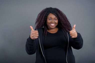 Gri arka planda izole edilmiş, olumlu içerik ve mutluluk duygularıyla baş parmaklarını gösteren, gündelik giysiler içindeki genç, iyimser Afro-Amerikan kadınına yakın çekim. Ortak alan, kalite ve tavsiye ile tatmin kavramı.