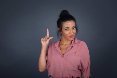 Bir parmağı havada, gri stüdyoda gülümseyen ve gülümseyen gündelik giysili çekici genç esmer kadın. 