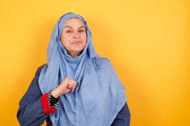 Tesettürlü neşeli, orta yaşlı Müslüman kadının yakın çekimi neşeli, memnun ve kendinden emin görünüyor, başparmağıyla işaret ediyor, stüdyo geçmişinde izole edilmiş..