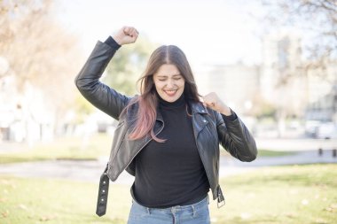 Genç, güzel, beyaz tenli esmer kadın. Deri ceket giyiyor. Bahar parkında bir şey kutluyor.