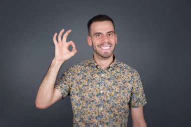 Çekici bir adamın işaret dilini iyi kullanmasına sevindim. Onayladığına, neşeli bir ifadeye sahip olduğuna. Yakışıklı erkek fotoğrafı çekici bir görünüme sahip, iyimser olmak. Gri duvara yaslanarak.