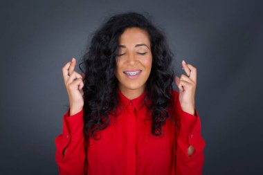 Kıvırcık saçlı, kırmızı tişörtlü neşeli esmer kadın dişlerini sıkıyor, parmaklarını çapraz yapıyor, arzu edilen dilekleri diliyor, gündelik kıyafetler giyiyor, iyi haberleri bekliyor, gri duvarın üzerinde duruyor.