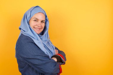 Orta yaşlı, neşeli, tesettürlü Müslüman bir kadının kolları bağlı bir şekilde evde duruşu. Kameraya bakıp gülümsüyor.. 