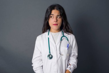 Şaşırmış genç beyaz kadın doktorun fotoğrafı dudaklarını kıvırıyor ve endişeli bir bakış atıyor. Önünde korkunç bir şey görüyor. Gri arka planda izole edilmiş, tıbbi üniforma giyiyor..