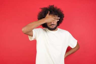 Afro saçlı genç adam gözlerini kırmızı duvara kapatıyor.   