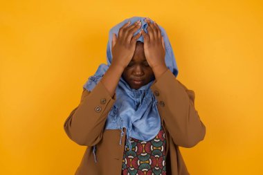 Güzel, Afro-Amerikan Müslüman bir kadın başını izole bir arka planda tutarak acı ve migrenden dolayı çaresiz ve stresli bir baş ağrısı çekiyor. Ellerini başının üstüne koy..