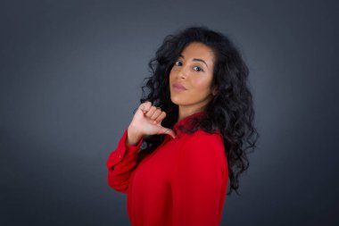 Kıvırcık saçlı, kırmızı tişörtlü neşeli, güzel esmer kadının yakın çekimi neşeli, memnun ve kendinden emin görünüyor. Başparmağıyla işaret ediyor, gri stüdyo arka planında izole edilmiş..