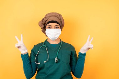 Arka planda iyimser bir gülümsemeyle izole edilmiş yeşil üniformalı güzel Müslüman kadın doktor portresi iki elinle de barış ya da zafer jesti gösteriyor, dost canlısı görünüyor. V işareti.