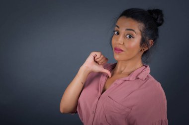 Neşeli, güzel, esmer bir kadının yakın çekimi neşeli, mutlu ve kendinden emin görünüyor. Başparmağıyla işaret ediyor. Gri stüdyo arka planında izole edilmiş..