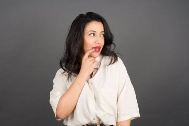 Güzel esmer kadın, sıradan giyinmiş, gözlerini kaçırıyor. Gri arkaplanda duruyor.  