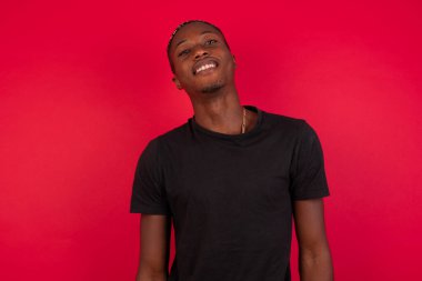 Afro-Amerikan adam, geniş gülümsemeli, beyaz dişli, sıradan tişörtlü, stüdyo duvarının başında duran, izinli olduğu için sevinen. İçerde dikilen adam.