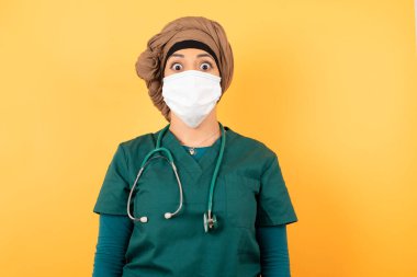 Şaşırmış, şaşkın bakışlı, şaşırmış, çığlık atan yeşil üniformalı, güzel, Müslüman bir kadın doktorun fotoğrafı. Şok olmuş bir şekilde kameraya bakıyor.