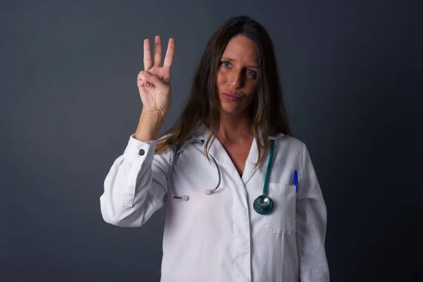 若いです医師の女性を身に着けています医療制服灰色の壁に立って表示し 指の番号3で指摘しながら 自信と幸せ — ストック写真