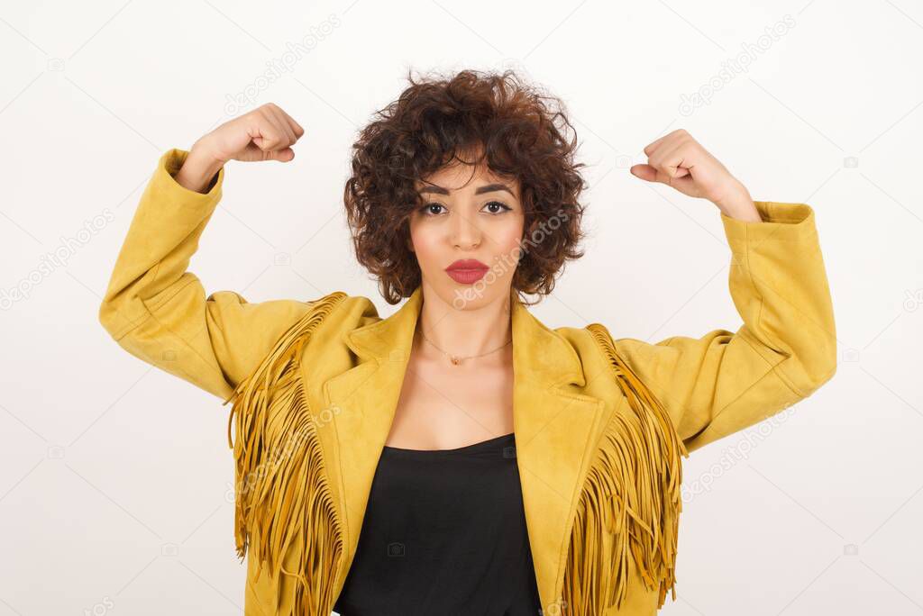 Waist up shot of woman raises arms to show her muscles feels confident in victory, looks strong and independent, smiles positively at camera, stands against gray background. Sport concept.