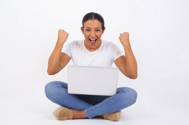 Sıradan tişörtler giyen, kollarını kaldıran, yumruklarını sıkan, sevinç ve heyecanla haykıran güçlü ve kararlı genç bir kadının portresi. Zafer, başarı ve başarı konsepti. Dizüstü bilgisayar kullanan güzel bir Arap kadın.