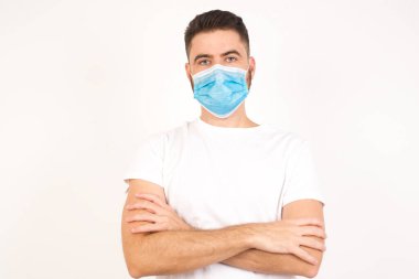Beyaz tenli, genç, yakışıklı bir erkeğin bel portresi pozitiftir. Kollarını kavuşturur, mutlu ve kendinden emin hisseder. Gri arka planda izole edilmiş günlük kıyafetler giyer. Tıbbi maske takan beyaz bir adam.