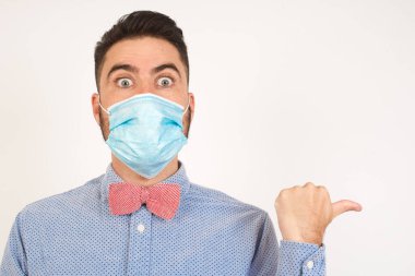 Baş parmağıyla tıbbi maske takan neşeli genç adamın yatay görüntüsü gri arka plan üzerinde izole edilmiş boş bir duvara işaret ediyor. Şuna bir bak. Etnik konsept.
