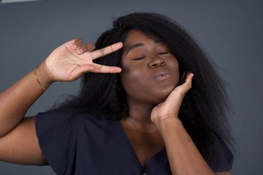 Güzel, güzel, Afro-Amerikalı bir kadın, gri duvarda dikilmiş, günlük giysiler giymiş, gözlerinin yanında V işareti yapıyor..