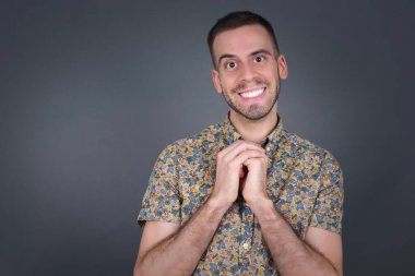 Tatlı yüzlü, rüya gibi, genç bir erkek, ellerini yüzünün yanında çapraz tutuyor, hoş bir şey için heyecanlı, gri arka plana karşı duruyor..