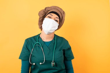 Genç, güzel, Müslüman doktor kadın yeşil üniformalı yüzünü saçlarıyla örterek kameraya bakıyor..