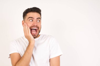 Şaşkınlık - adam heyecanla yüzüne bakıyor. Heyecandan yana bakan mutlu genç adam şaşırdı. Gri arka planda beyaz tişört giyen beyaz bir genç..