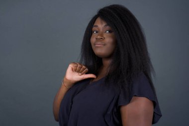 Neşeli, güzel, Afro-Amerikan kadının yakın çekimi neşeli, memnun ve kendinden emin görünüyor. Başparmağıyla işaret ediyor. Gri stüdyo arka planında izole edilmiş..