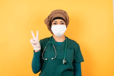 Yeşil üniformalı genç, Müslüman doktor kadın duvarın karşısında duruyor ve parmaklarını iki ya da Ziyaretçi işaretiyle gösterirken kendinden emin ve mutlu gülümsüyor..