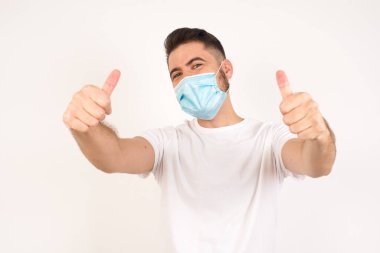 Sarışın genç bir adam tıbbi maske takıyor ve izole edilmiş bir geçmişi var. Elle olumlu jest yapıyor, gülümsüyor ve başarı için mutlu. Kameraya bak, kazanan jest..