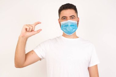 Tıbbi maske takan sarışın bir adam izole edilmiş arka plan jestleri ile küçük boy, ölçü sembolü gösteriyor. Kameraya bakıp gülümsüyor. Ölçüm konsepti.