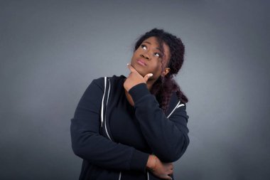 Düşünceli gülümseyen Afro-Amerikan kadın portresi elini çenesinin altında tutuyor, yan bakıyor, ilgi çekici bir şey düşünüyor ya da merak ediyor, sıradan giyiniyor, gri stüdyoya karşı poz veriyor. Karar alma kavramı.