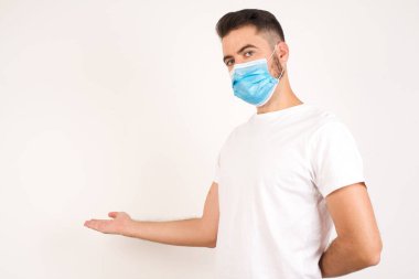 Sağlık maskesi takmış yakışıklı bir gencin portresi. Gri arka planda hoş geldin jesti yapıyor..