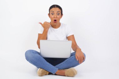 Genç ve neşeli bir kadının başparmak uçlarında dizüstü bilgisayarla çekilmiş yatay görüntüsü beyaz arka planda izole edilmiş boş bir duvara işaret ediyor. Şuna bir bak. Etnik konsept.