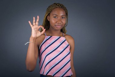 Çekici, Afro-Amerikalı genç bir kadının el işareti göstermesine sevindim. Onayladığının göstergesi olarak, neşeli bir ifadesi var. Gri duvara yaslanarak.