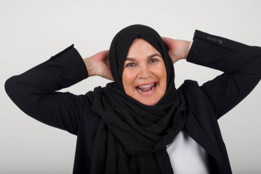 Güven ve cinsel ilişki konsepti. Siyah tesettürlü genç Müslüman bir kadının portresi. Kendine güvenen bir ifadeyle gülümsüyor ve ellerini başının üstüne koyuyor. Gri arkaplanda duruyor.