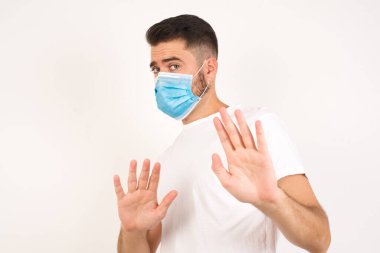 Tecrit edilmiş arka planda tıbbi maske takan yakışıklı genç adam korkmuş, korkmuş ve tiksinmiş ifade iki elinle hareket etmeyi kes ve orada kal de. Panik konsepti.