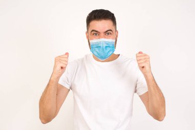 Sıradan tişörtler giyen, kollarını kaldıran, yumruklarını sıkan, sevinç ve heyecanla haykıran güçlü ve kararlı genç bir erkeğin portresi. Zafer, başarı ve başarı konsepti. Tıbbi maske takan beyaz bir adam. 