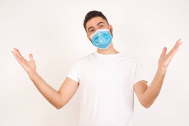 Tıbbi maske takan neşeli adam beyaz arka planda izole edilmiş kollarını kaldırarak hoş geldin jesti yapıyor..
