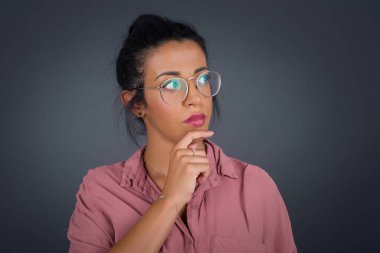Gözlüklü genç kadın elini çenesinin altında tutuyor, düşünceli bir şekilde yukarı bakıyor, gri arka planda duruyor ve mesajın için boş yer var. 
