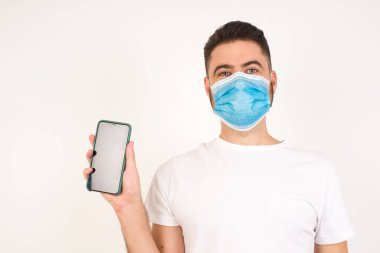 Stüdyoda tıbbi maske takan genç bir adam izole edilmiş arka planda akıllı telefon kullanıyor. 