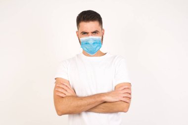 Beyaz arka planda izole bir şekilde duran, tıbbi maske takan ve kollarını kavuşturan kızgın, yakışıklı bir beyaz adam resmi. Hayal kırıklığına uğramış bir ifadeyle kameraya bakmak.