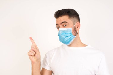 İşaret vermeyen bir adam. Yakın plan portre mutsuz, ciddi bir beyaz adam tıbbi maske takıyor ve parmağını kaldırıyor. Beyaz arka planda duruyor. Olumsuz duygular, yüz ifadeleri, duygular.